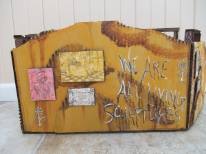 Back view, depicted as a wall in an abandoned house complete with skeletal themed artwork and wallpaper print, "We are all living sculptures"
